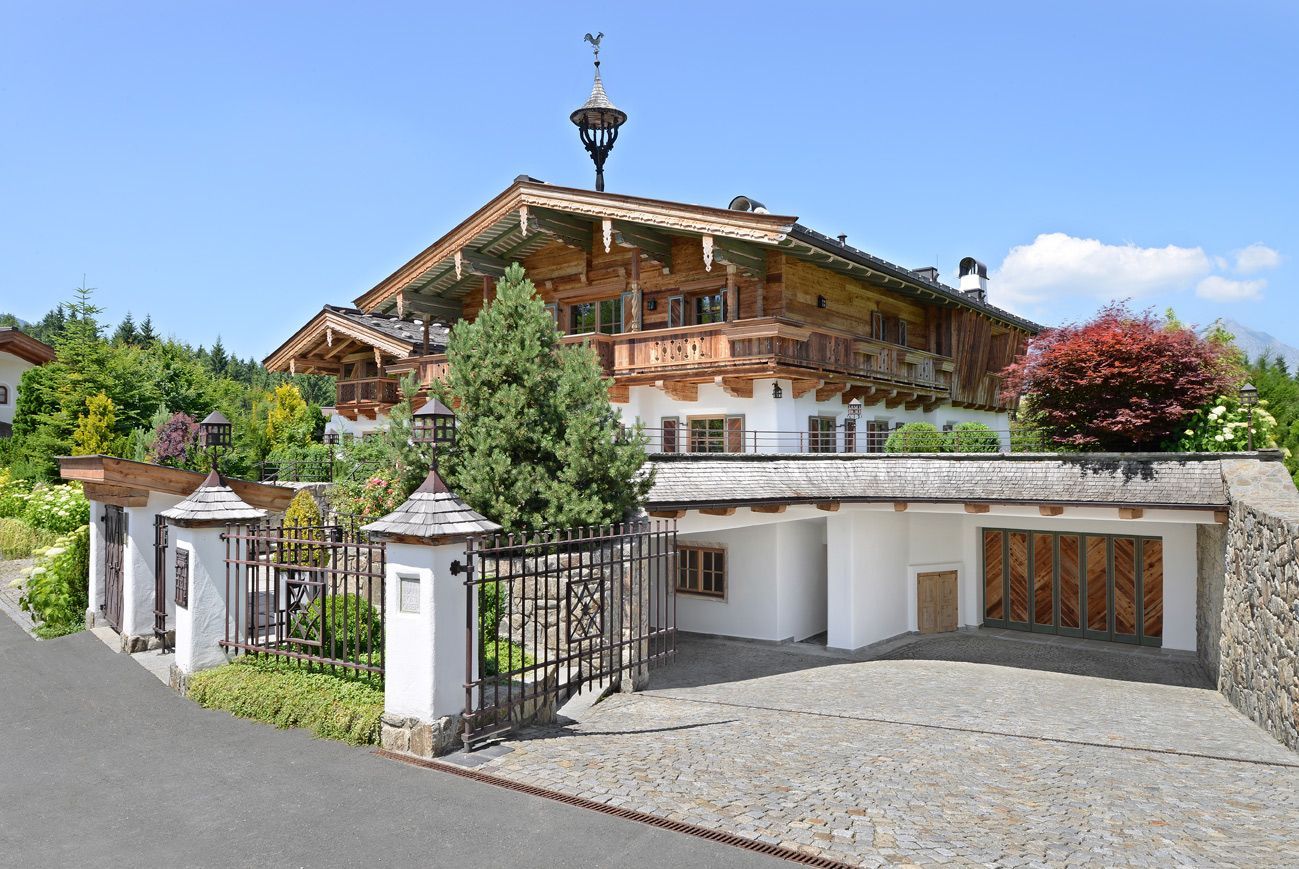 Elegant alpine-style house with carved wooden balconies, gated entrance, and a two-car garage under a blue sky.