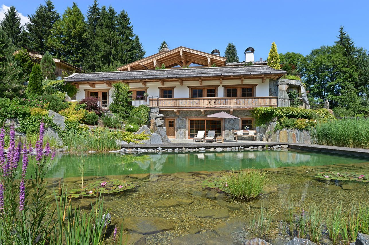 Luxury chalet by a pond, surrounded by lush vegetation and rocky landscape, blue sky above.
