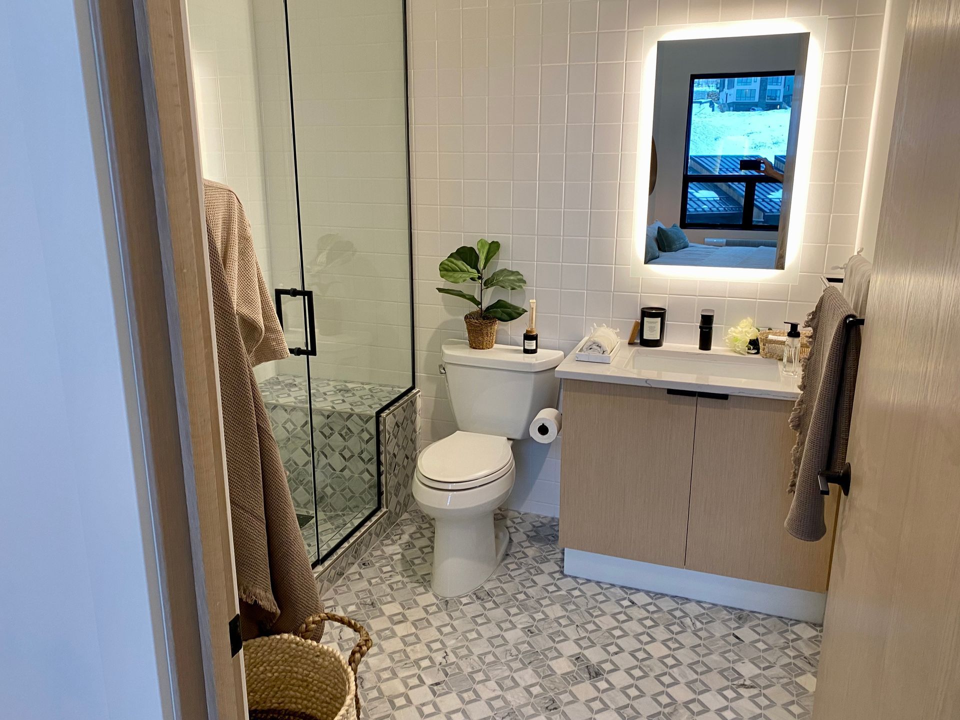 A modern bathroom with a glass shower, toilet, light-up mirror, and light wood vanity.