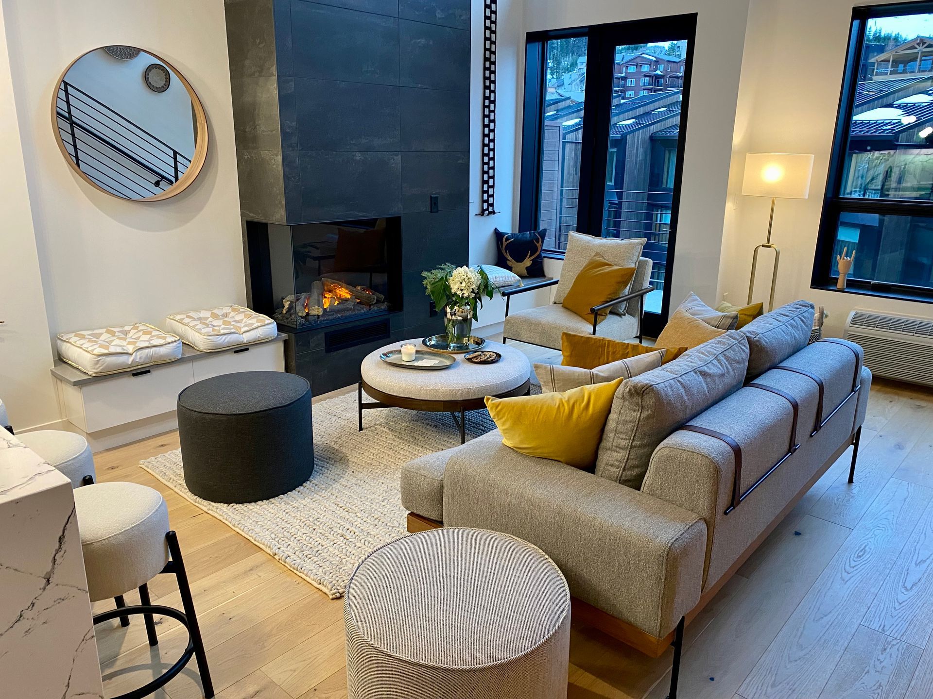Cozy living room with fireplace, sofa, and city view through windows. Furnished with round ottoman and rug.