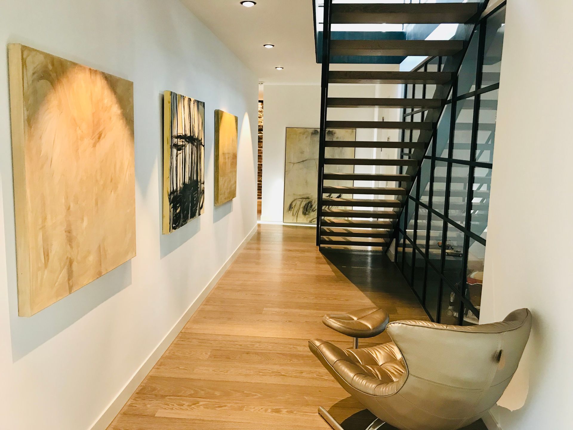 Hallway with art, wood flooring, and metal staircase leading upstairs. Gold chairs.