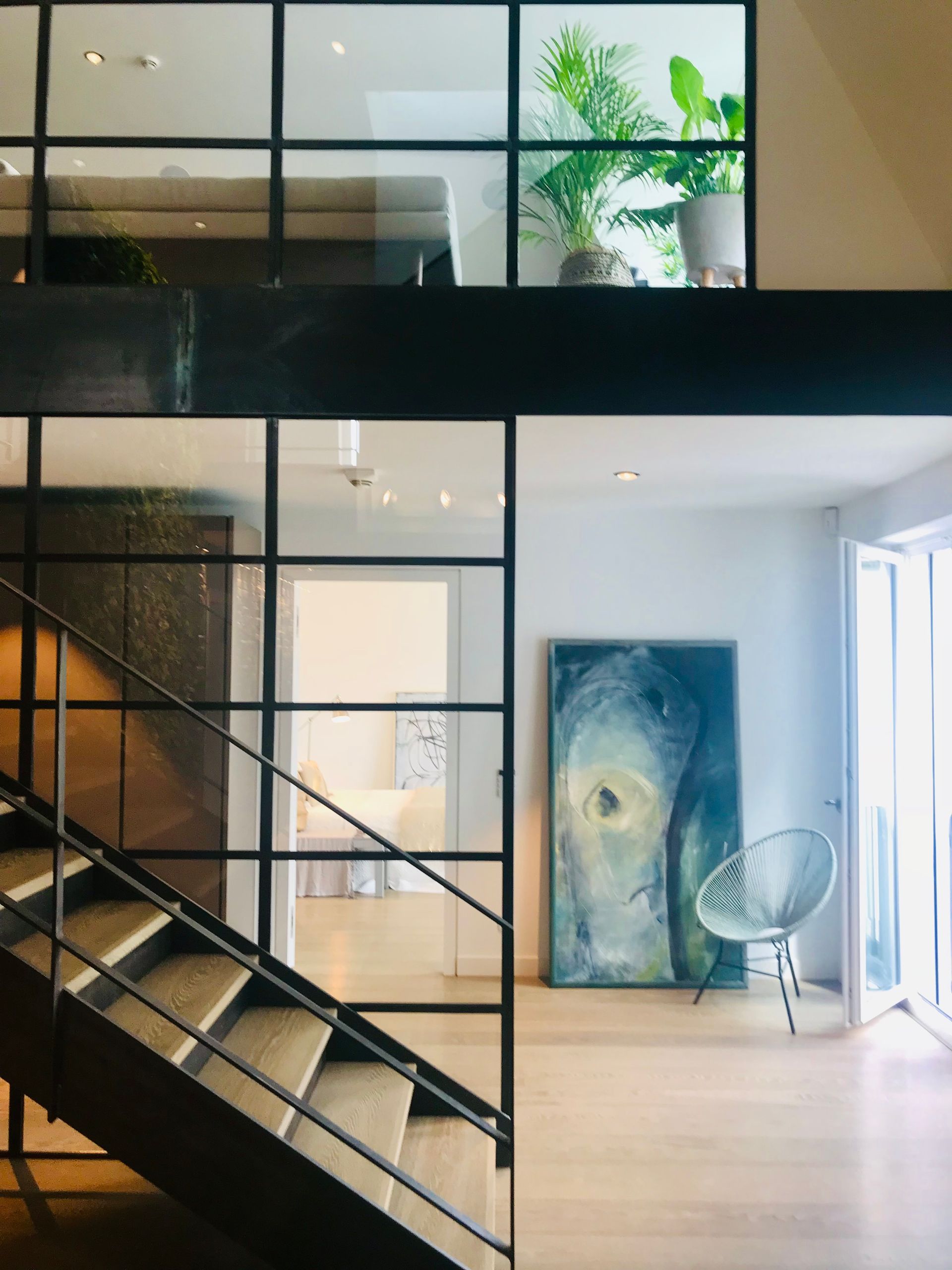Modern interior with staircase, steel-framed glass walls, artwork, chair, and plants. Bright, airy space.