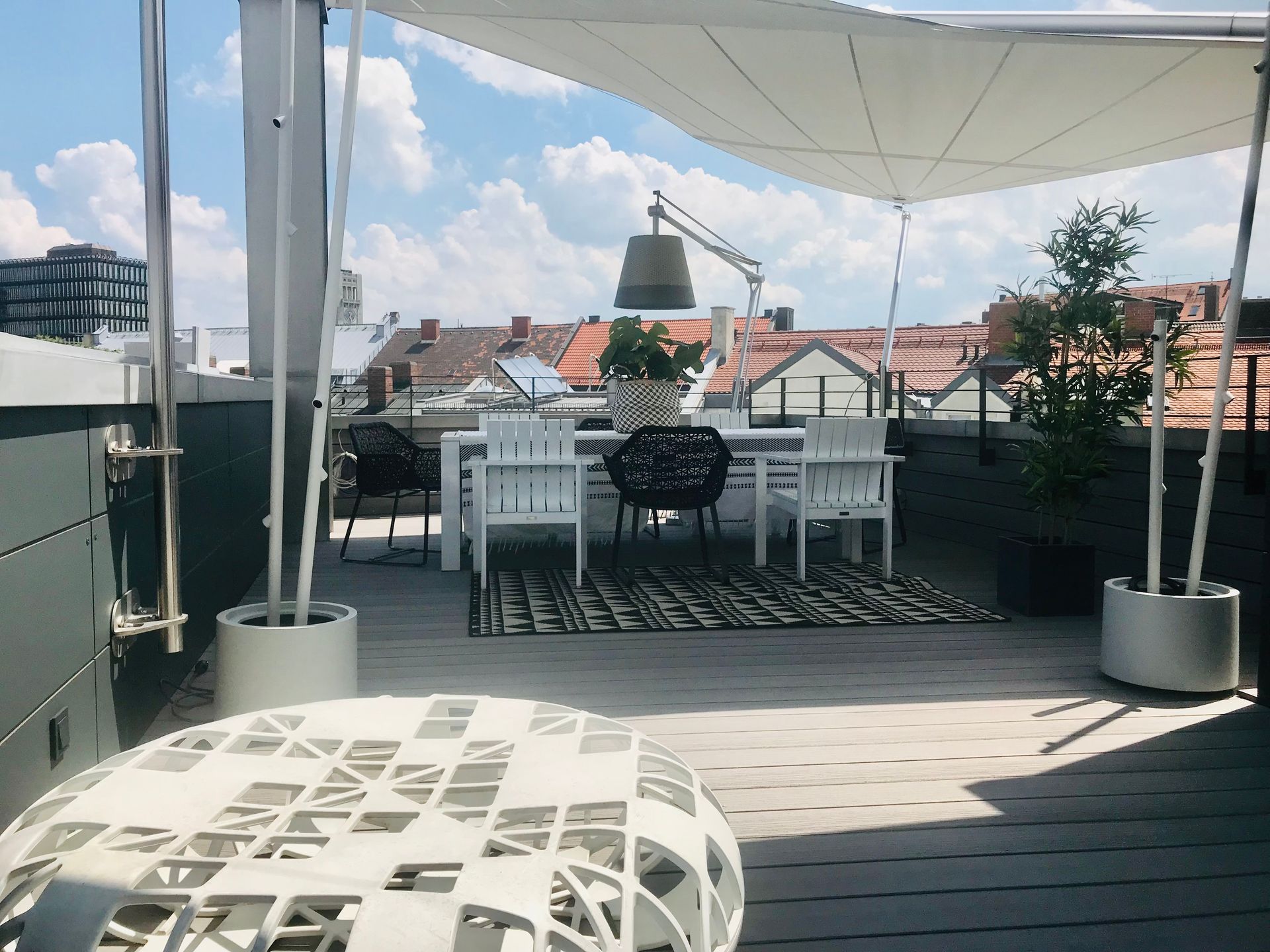 Rooftop patio with table, chairs, plants, and awning, overlooking a city.