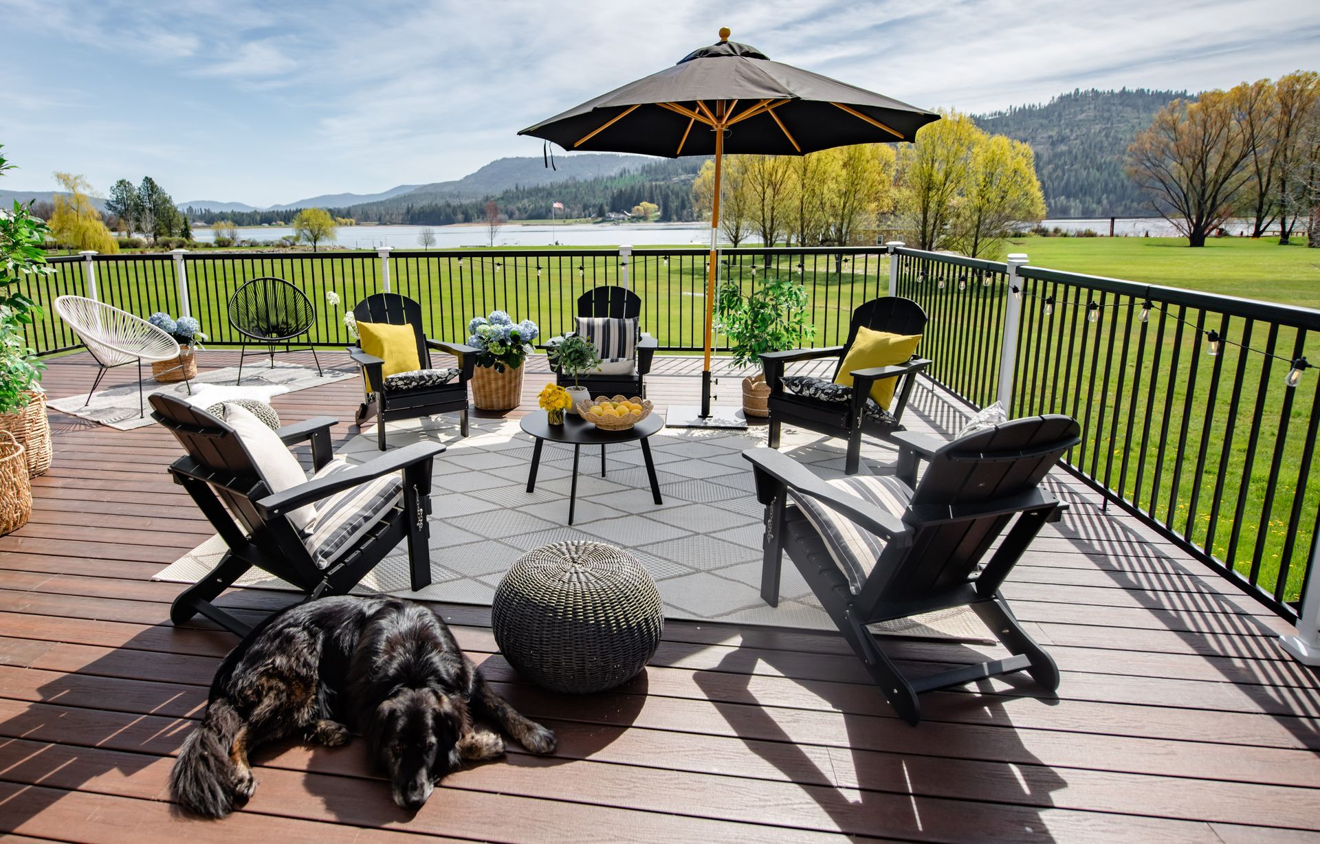 Backyard deck with black chairs, umbrella, fire pit, and dog, overlooking a green lawn and hills