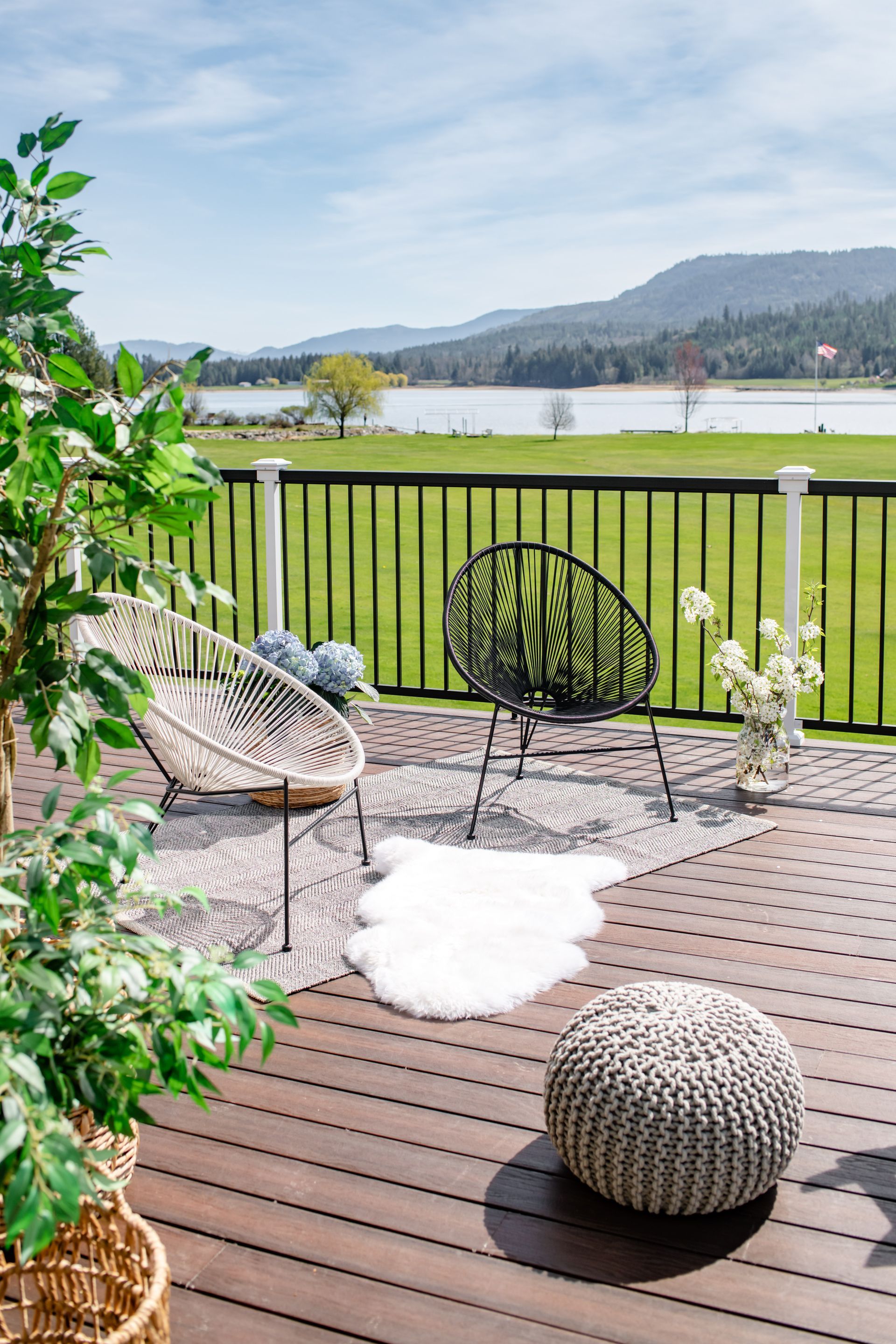 Patio with wicker chair, fluffy rug, round stool, and black railing overlooking a grassy lake and hills