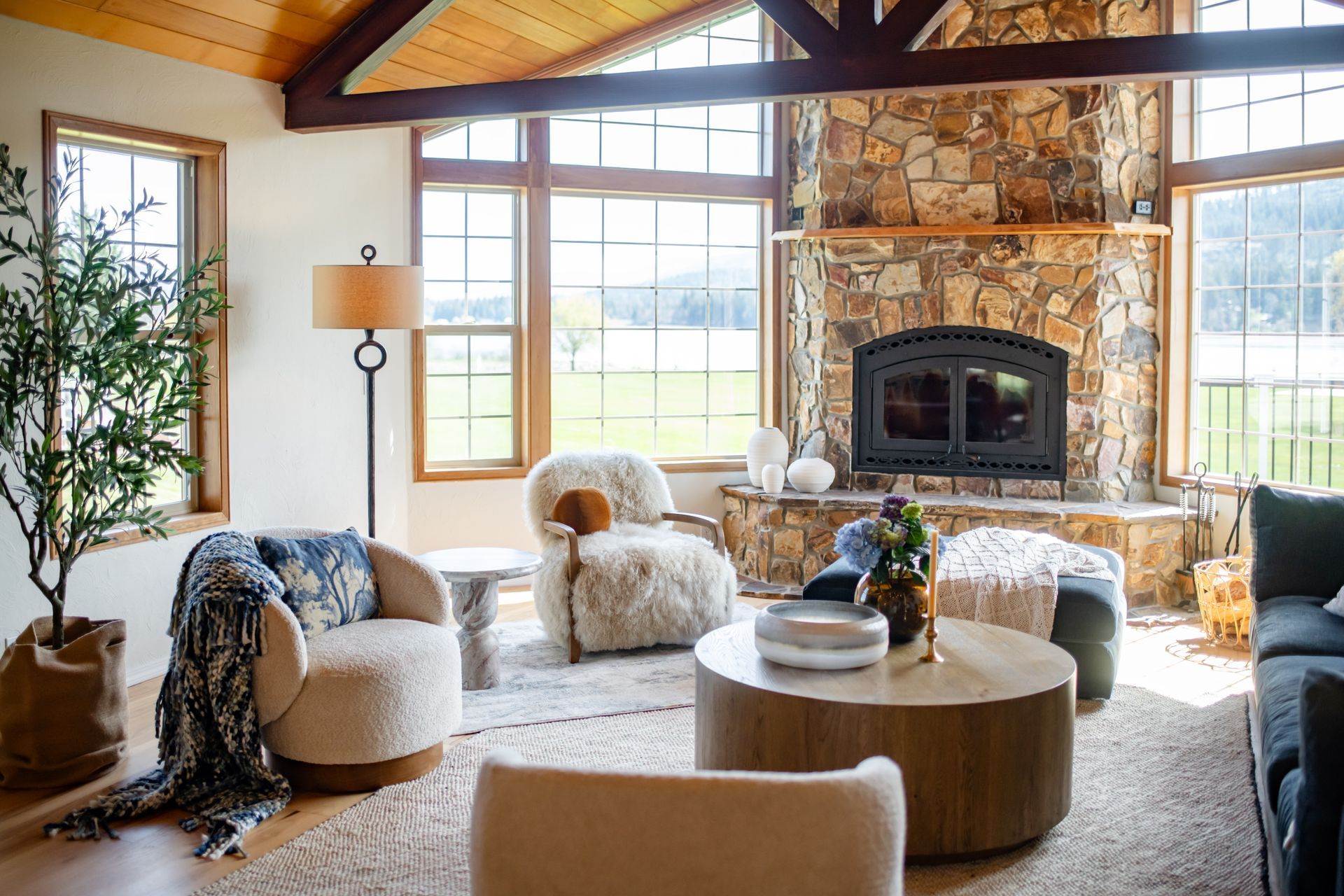 Bright living room with stone fireplace, large windows, and cozy cream chairs around a round wooden table