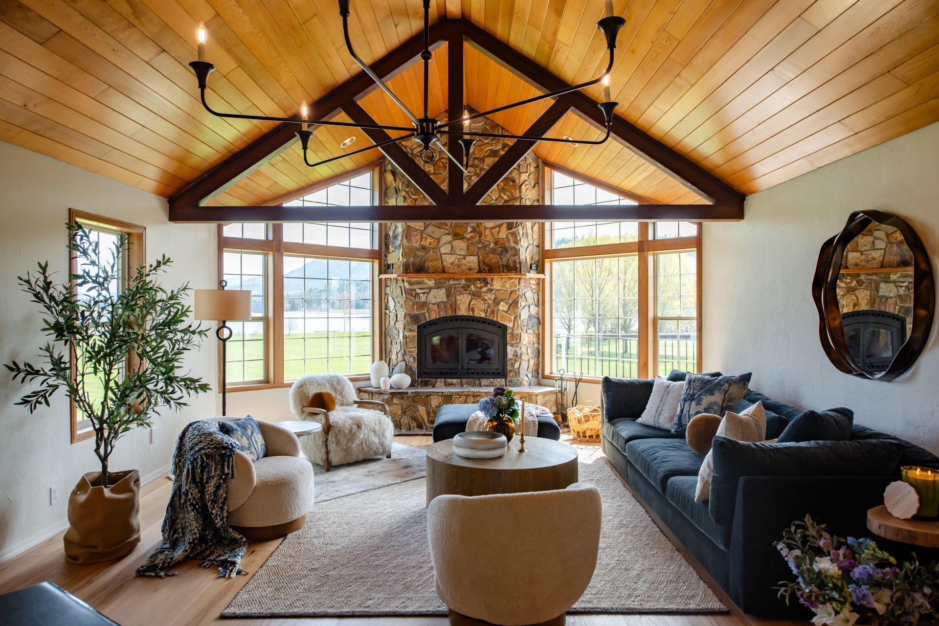 Cozy living room with vaulted wood ceiling, stone fireplace, large windows, and dark sofa seating.
