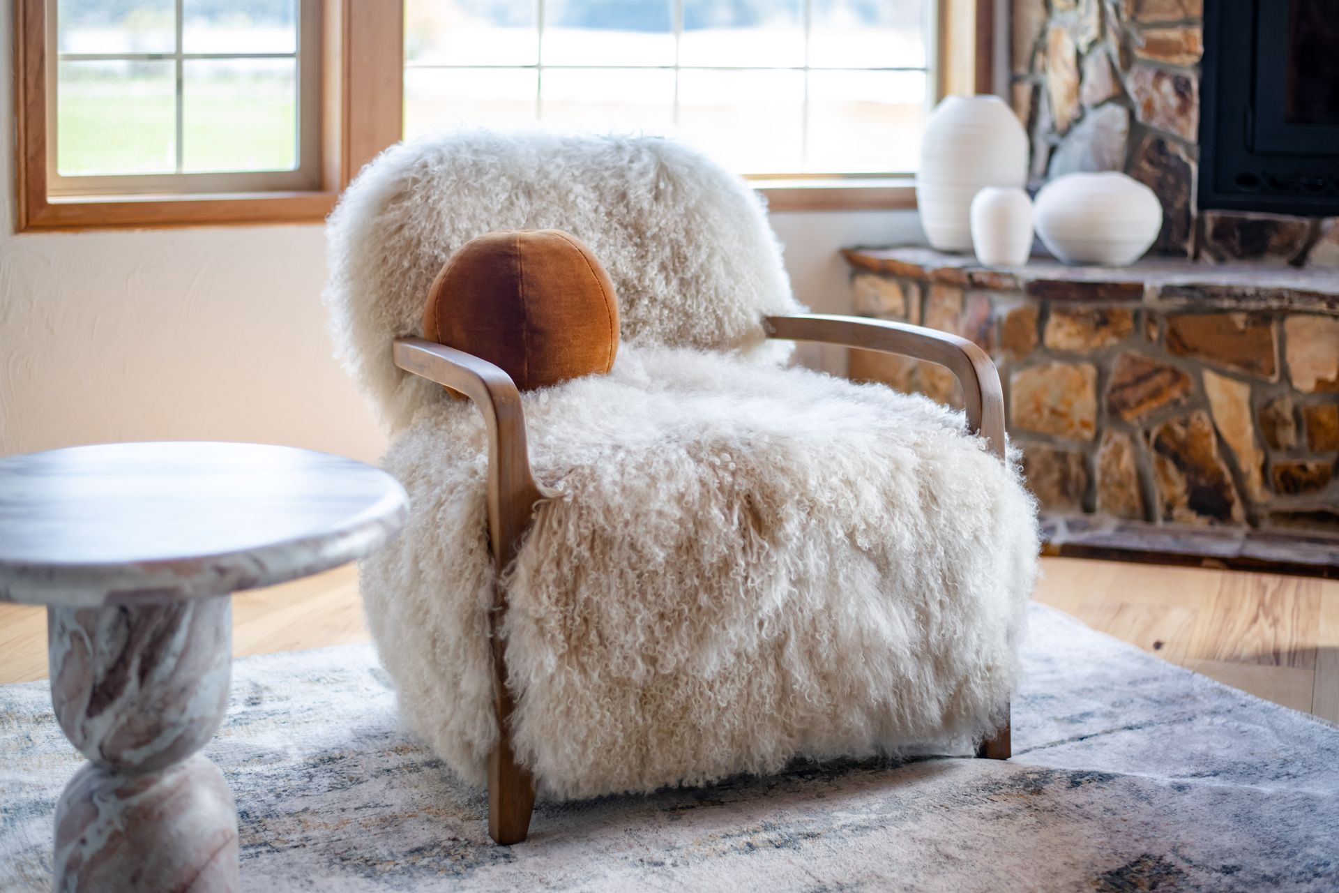 Cozy armchair with fluffy white shearling in a sunlit living room beside a rustic stone fireplace