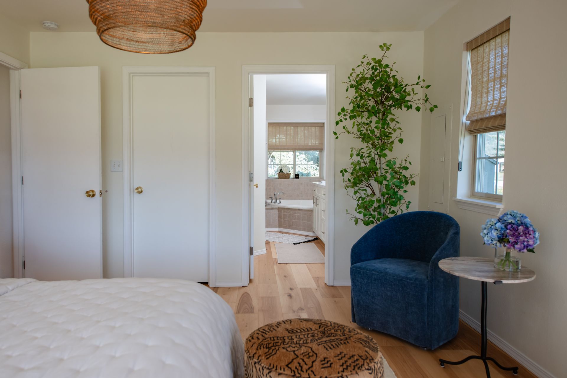 Bright bedroom with white walls, a blue chair, woven rug, potted plant, and doorway to another room