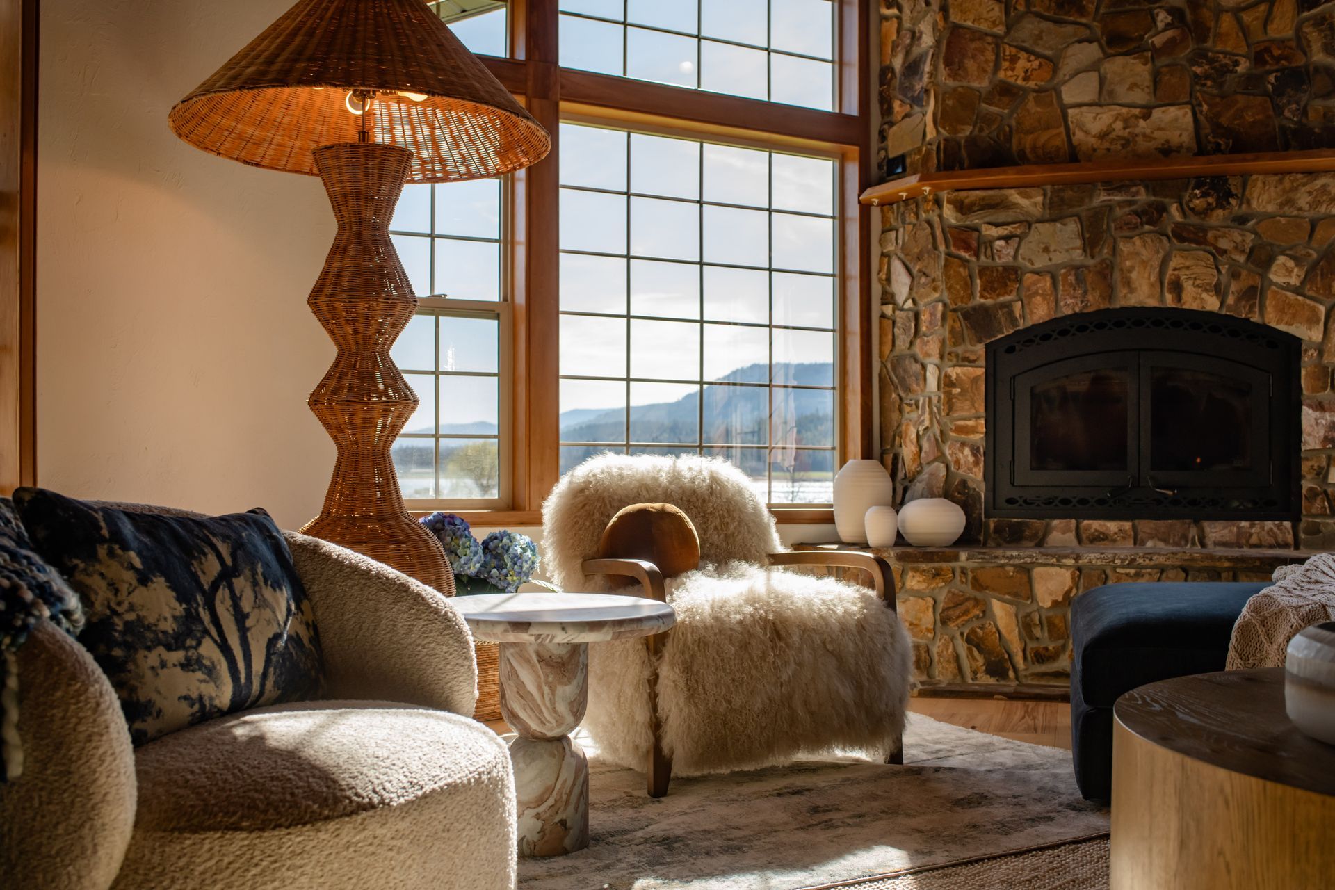 Cozy rustic living room with stone fireplace, large windows, and plush beige seating in warm sunlight