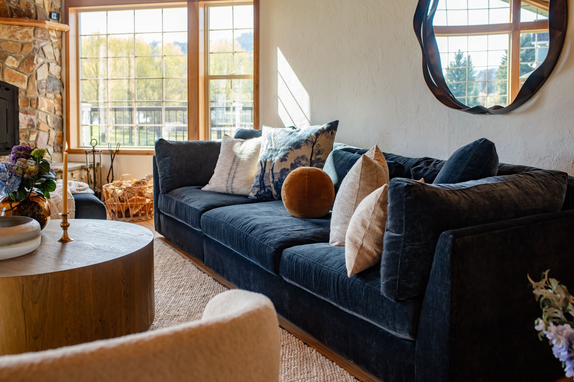 Cozy living room with a dark blue sofa, round wooden table, large windows, and a wall mirror