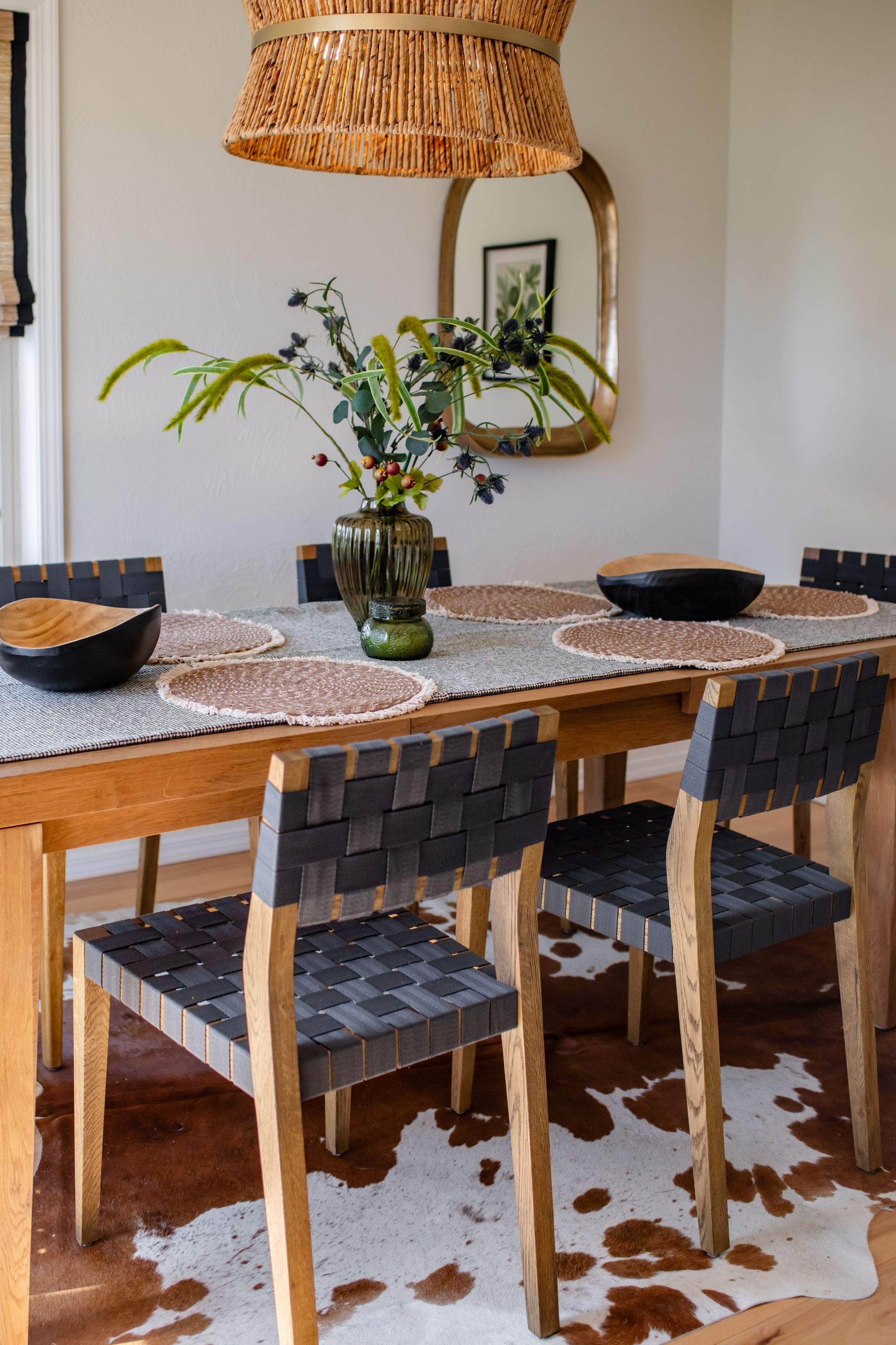 Dining table with woven chairs, patterned rug, vase of flowers, and woven pendant light in a bright room