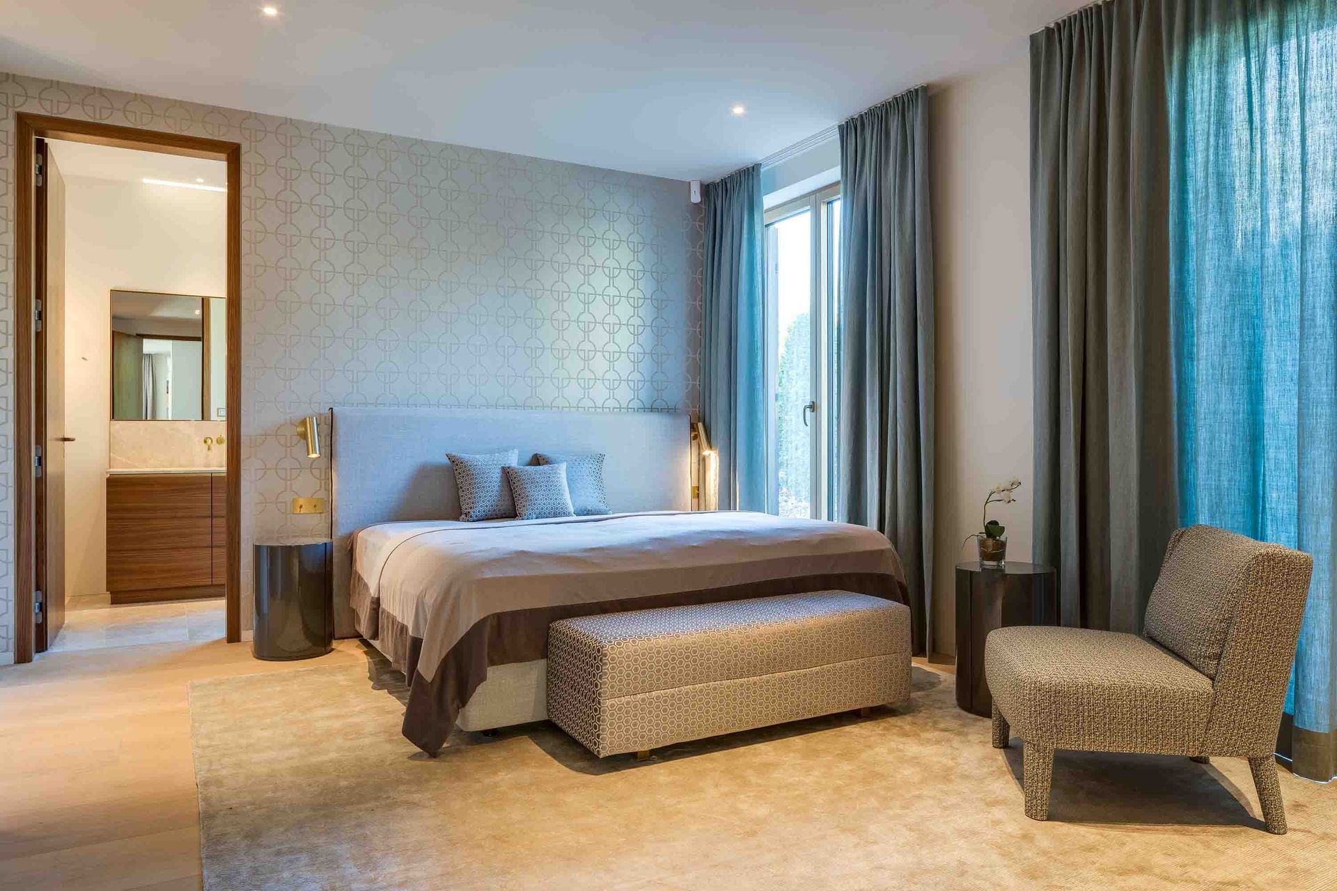 Bedroom with bed, bench, chair, and large windows with gray curtains. Light gray walls. Door to bathroom.