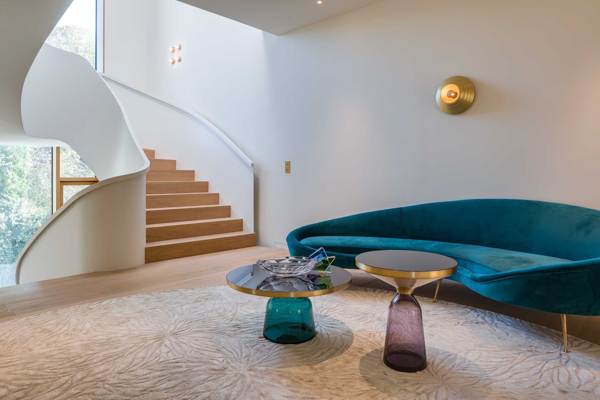 Teal curved sofa, two glass tables, and spiral staircase in modern living room.