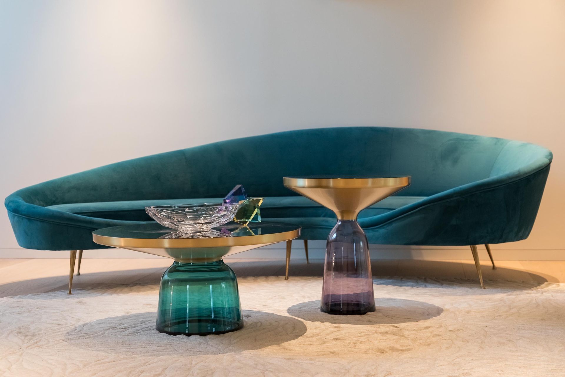Teal velvet sofa with gold legs, two glass tables, and a white rug.