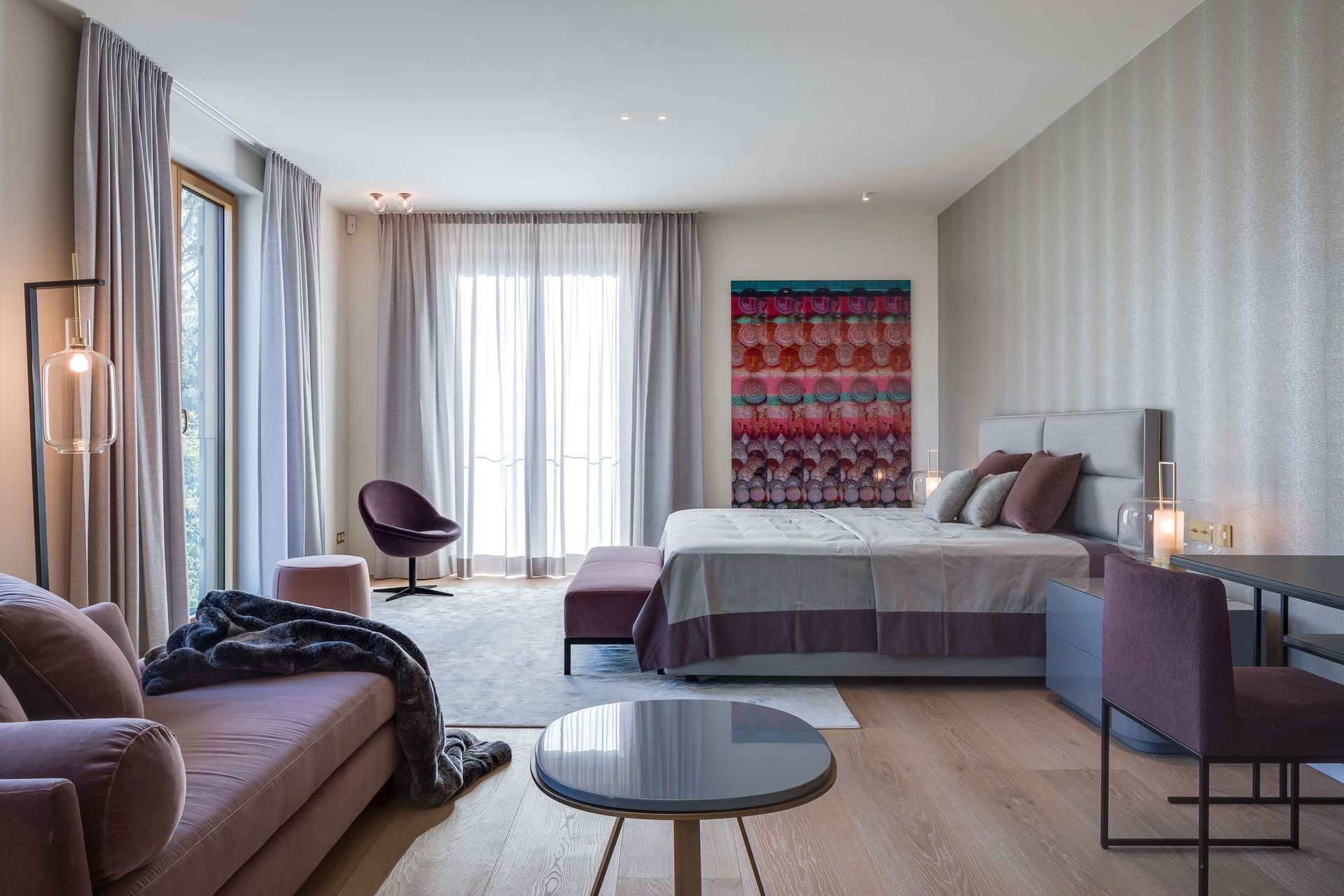 Spacious bedroom with light-colored walls, large windows, and modern furniture in shades of purple and gray.