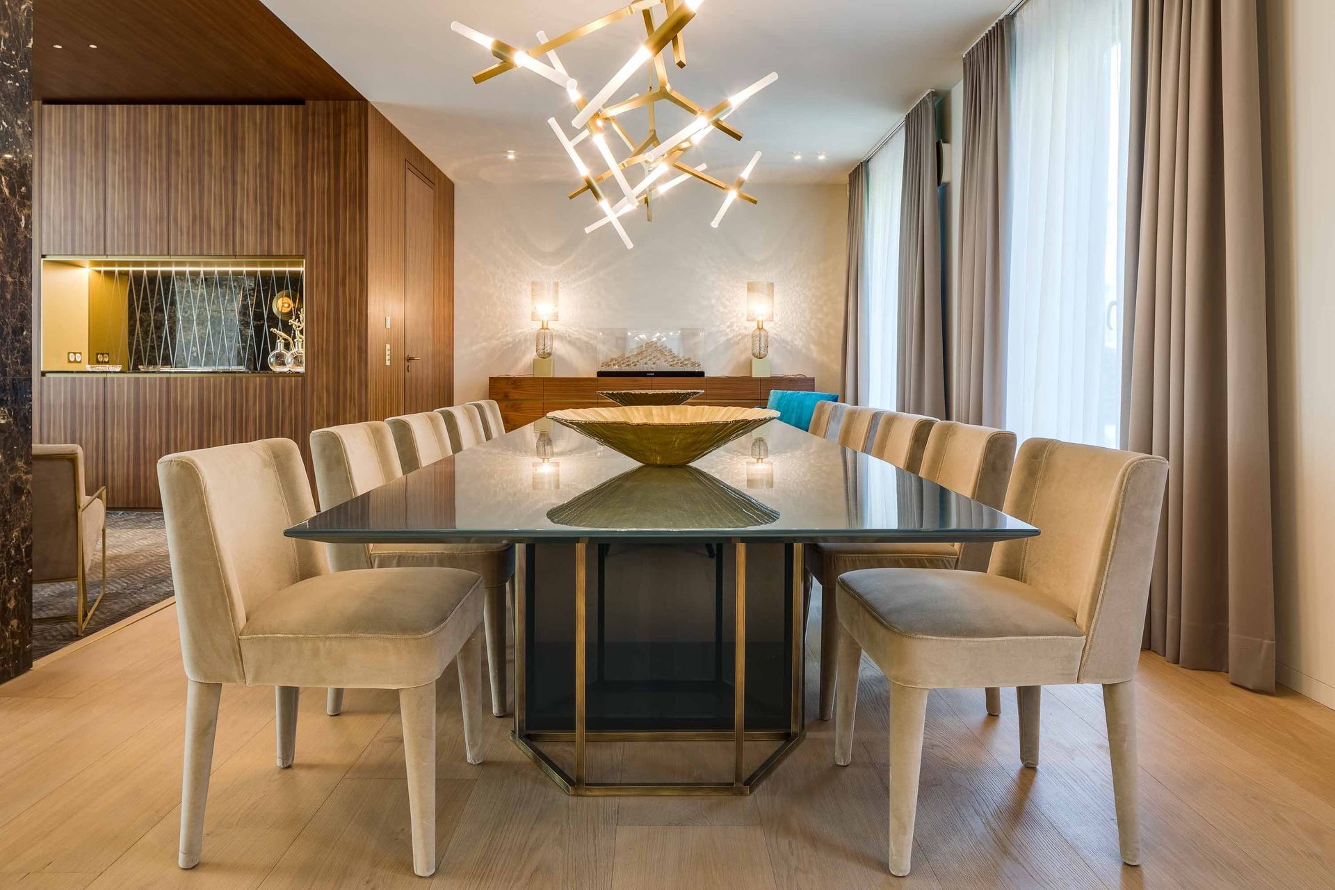 Dining room with a long table, chairs, and chandelier. Wooden paneling and curtains are also visible.