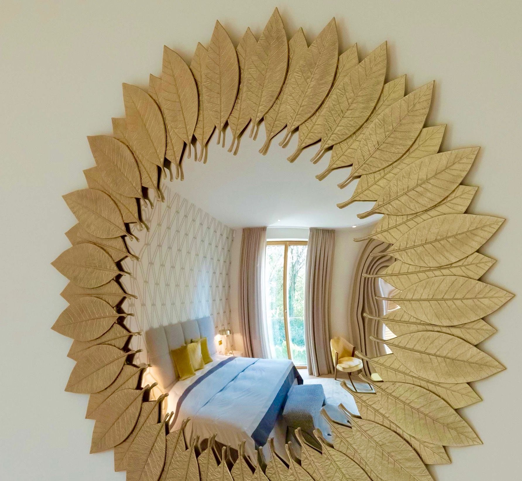 Gold leaf-framed mirror reflecting a bedroom with a bed, curtains, and a chair.