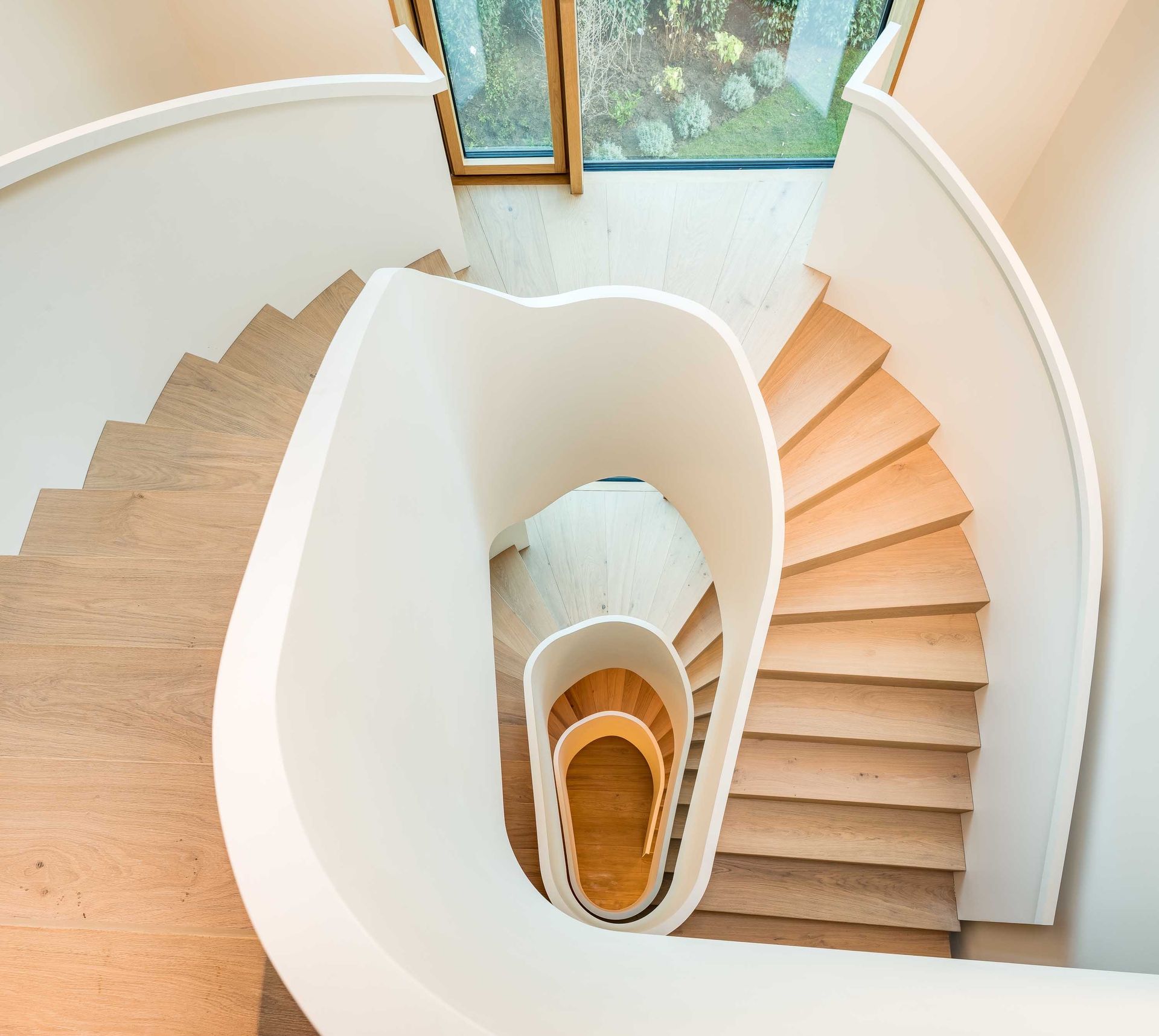 Bird's-eye view of a curved staircase with a white railing and wooden steps, leading up to a window.