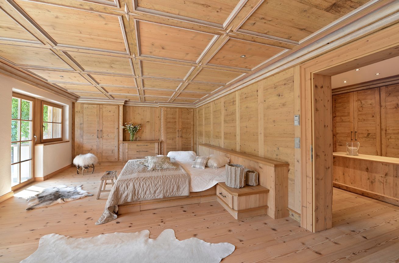 Wooden-paneled bedroom with bed, nightstand, ceiling, walls, and doorway to an adjacent room; natural light streams in.