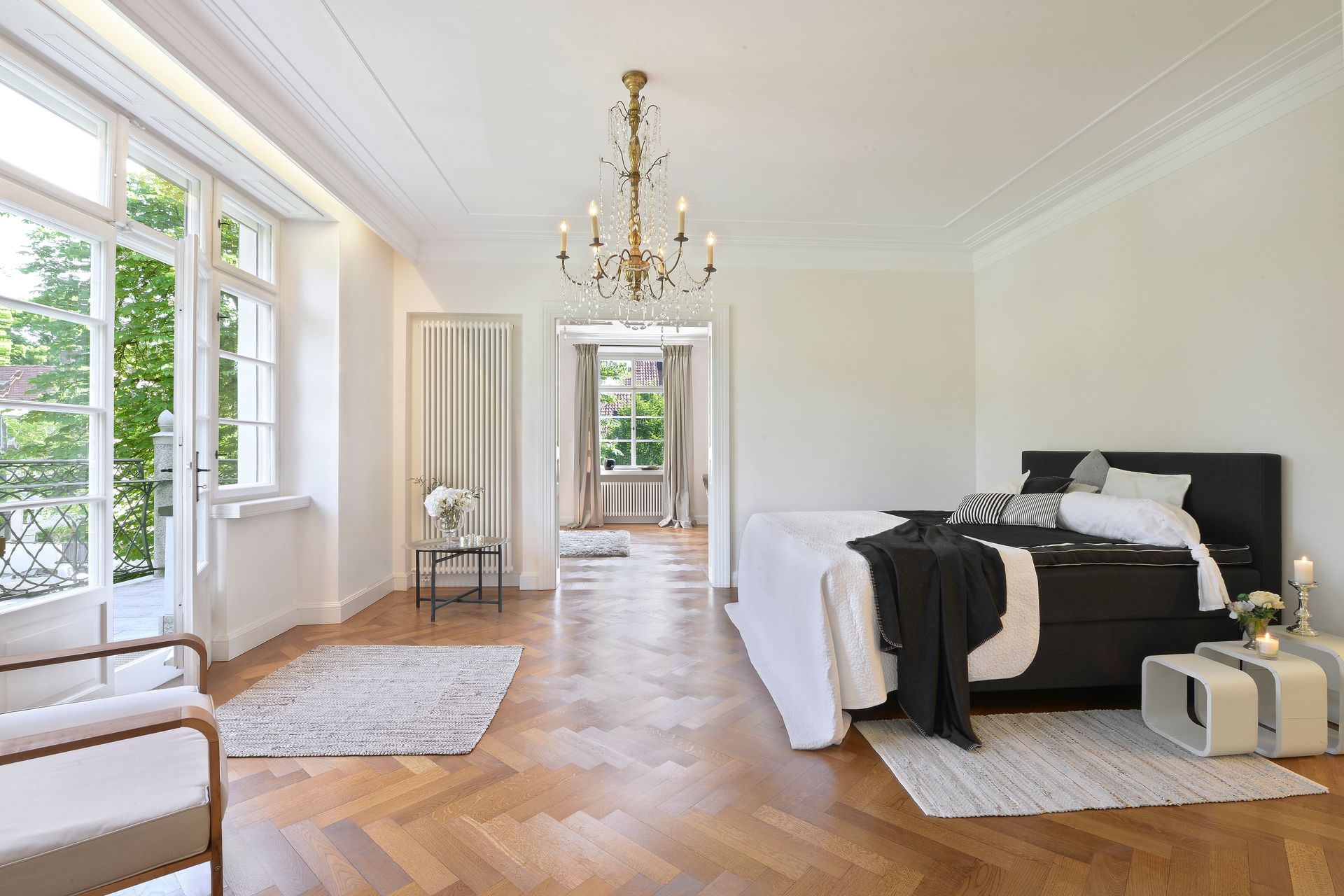 Bedroom with large windows, chandelier, hardwood floors, bed with black headboard, white bedding.