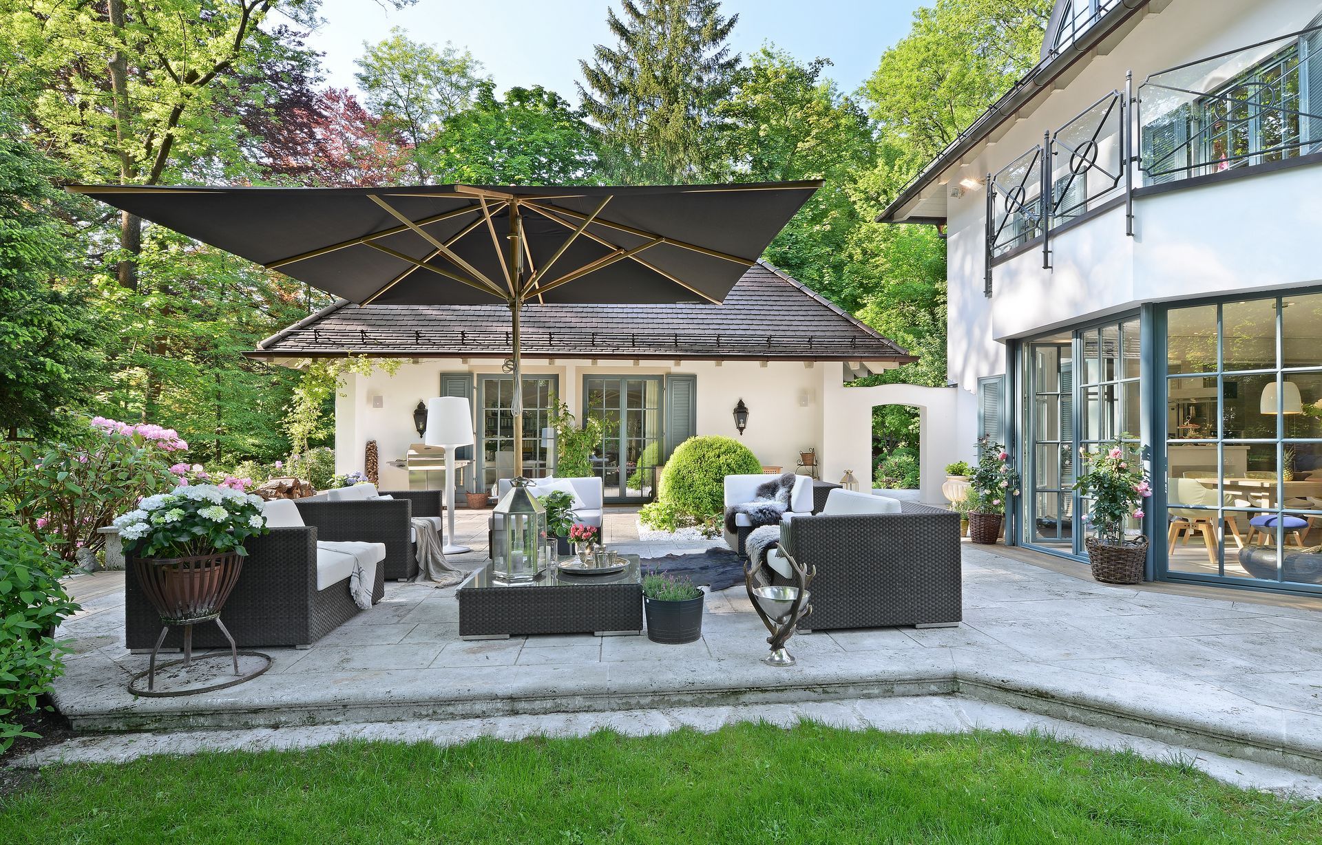 Outdoor patio with black umbrella, seating area, white building, and greenery.