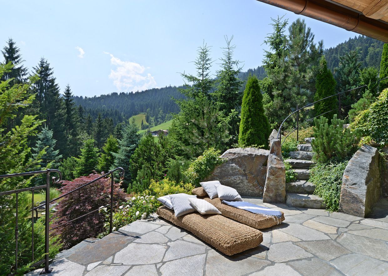 Two lounge chairs on a stone patio overlooking a mountain forest on a sunny day.
