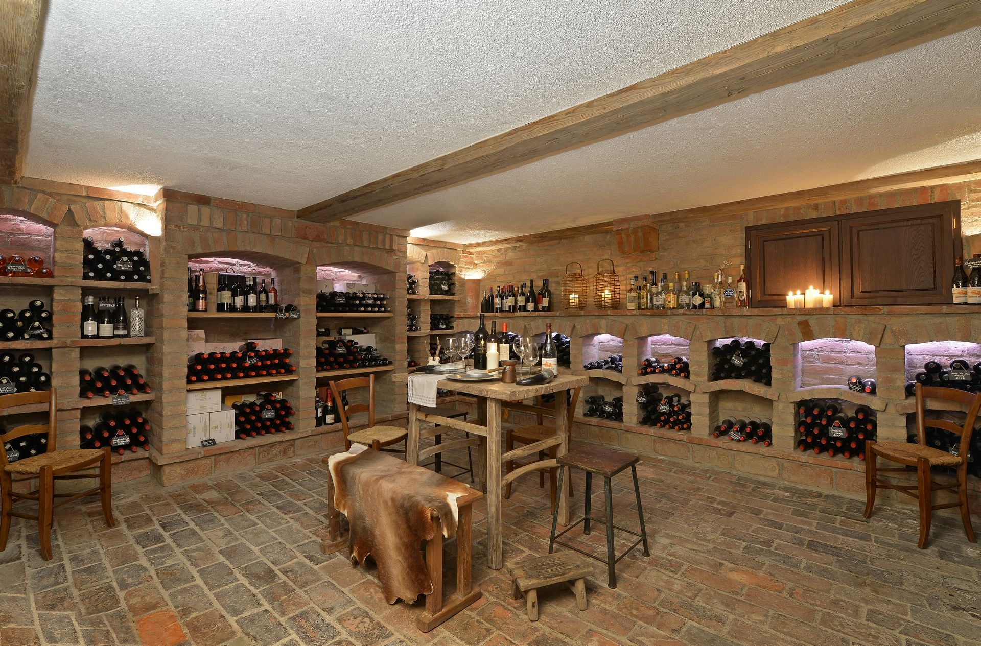 Wine cellar with brick walls and shelves filled with bottles; a table and chairs are in the center.