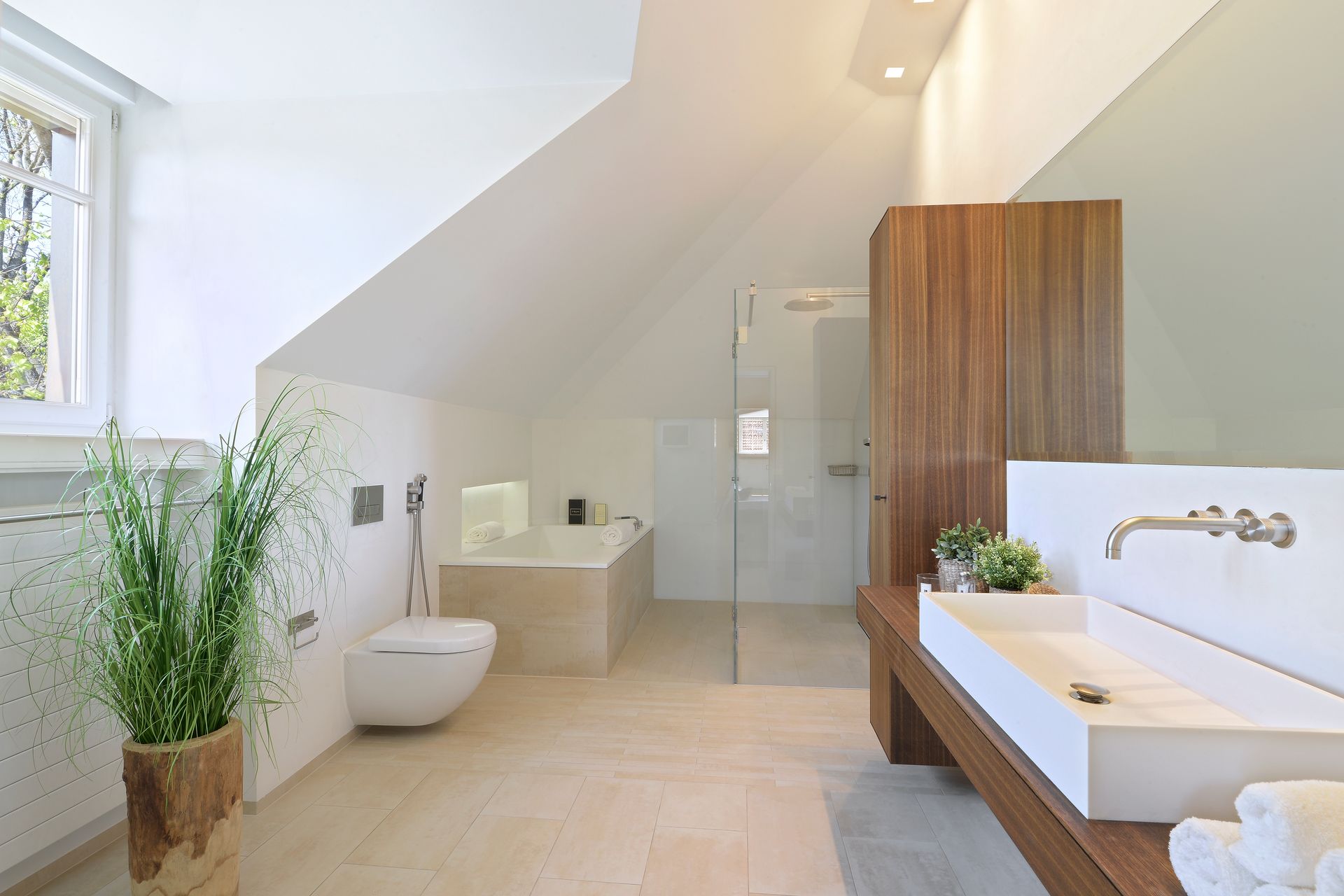 Modern bathroom with slanted ceiling, large sink, wooden cabinet, and plants.