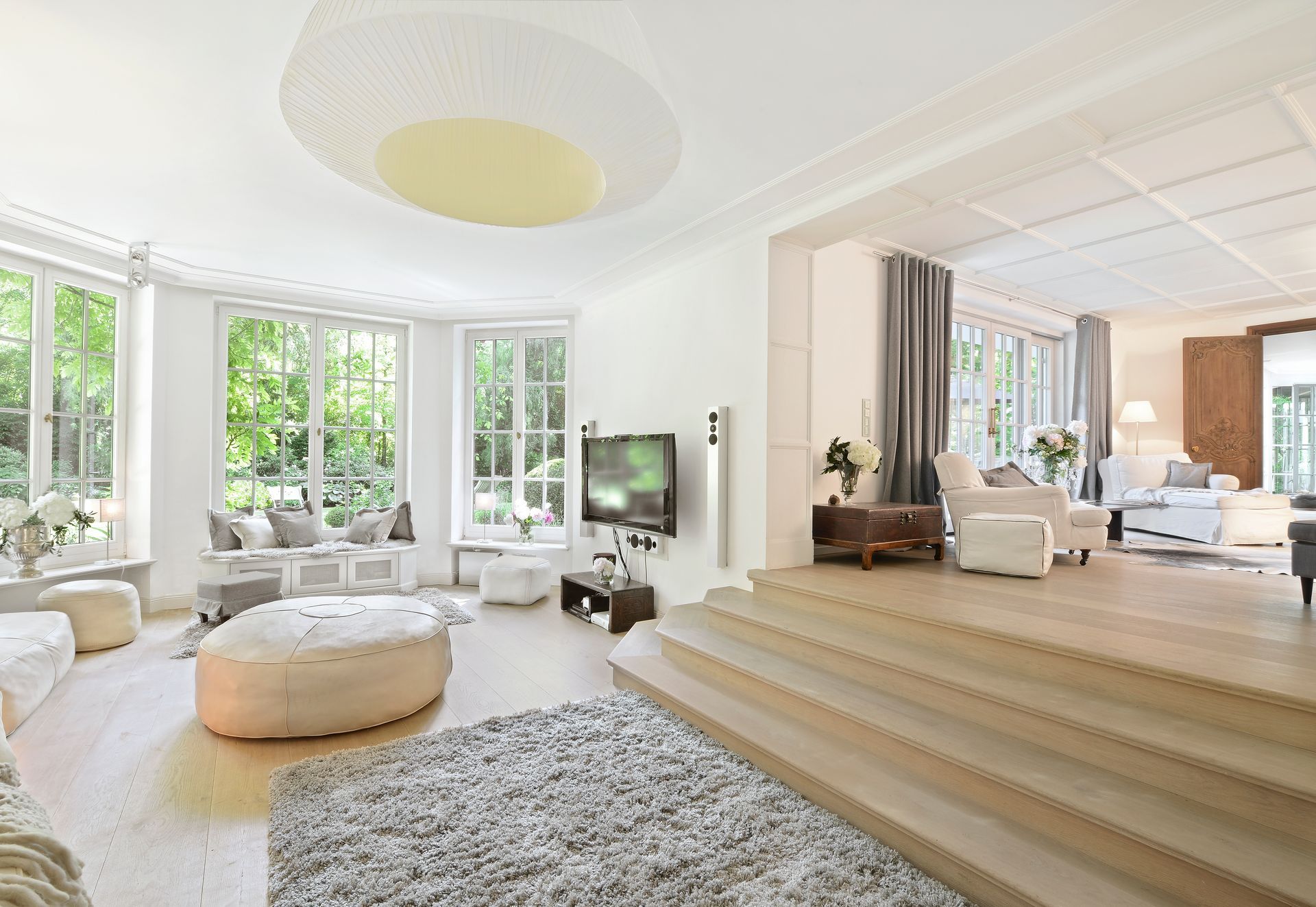 Bright living room with large windows, light-colored furniture, and a rug. Steps lead to a seating area.