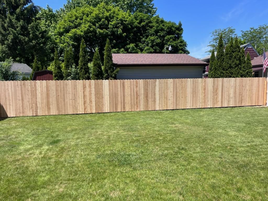 Wooden fence in a backyard with green grass, a garage, and trees under a blue sky.