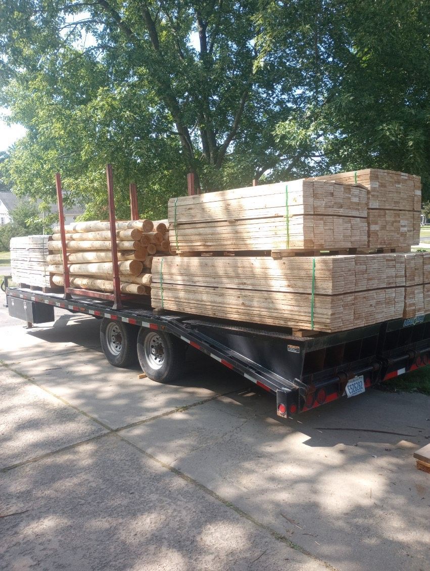 Flatbed truck loaded with stacks of lumber on a paved driveway in front of a tree.