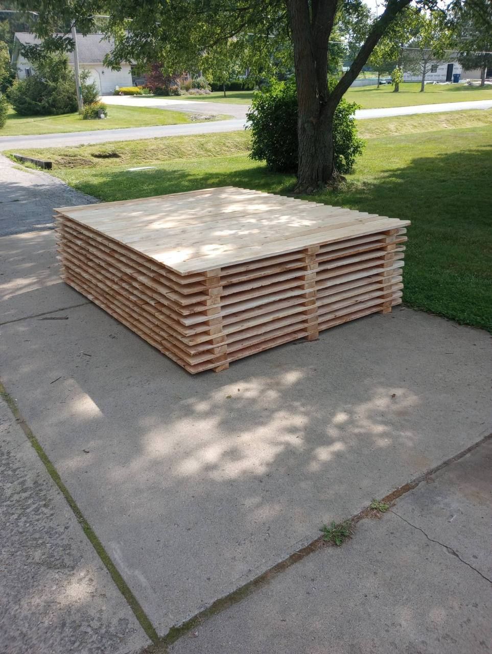 Stack of wooden boards on a concrete driveway, near a tree and residential area.