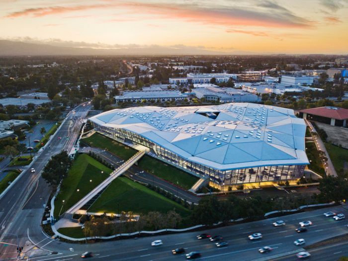 Aerial view of a large modern sports stadium with a white roof at sunset, beside busy roads and parking lots