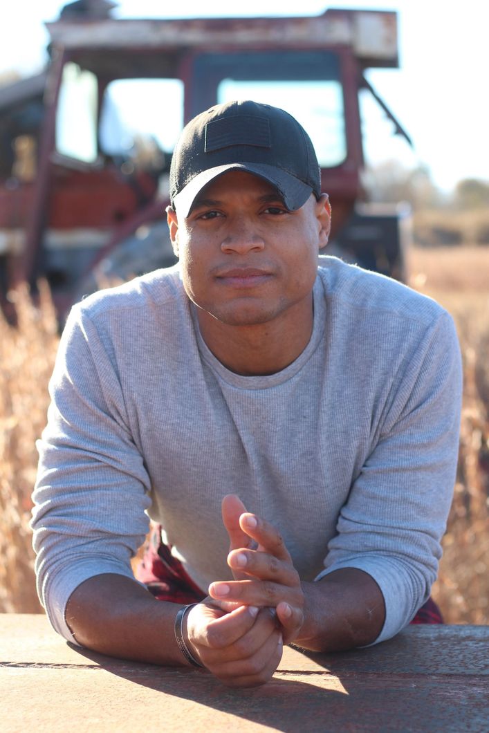 A man in a gray shirt is standing in a field with mountains in the background.