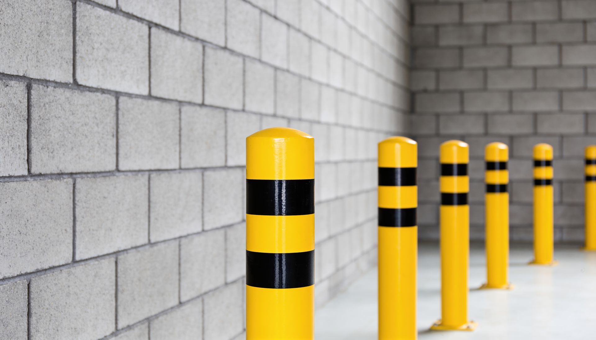 Row of bright yellow safety bollards with black stripes lining a gray concrete block interior wall.