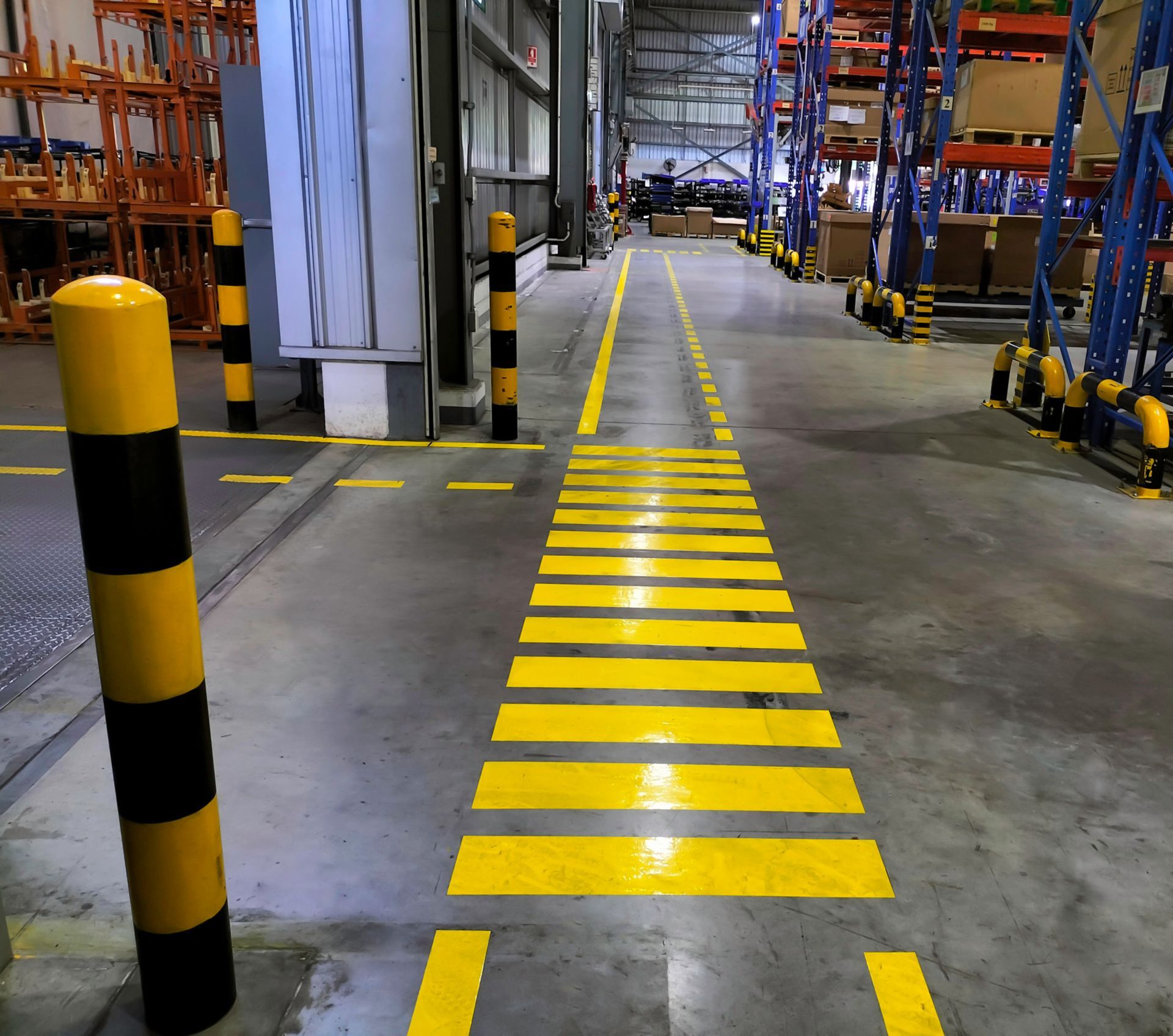 Bright yellow floor striping runs past striped safety bollards toward rows of tall pallet racking.