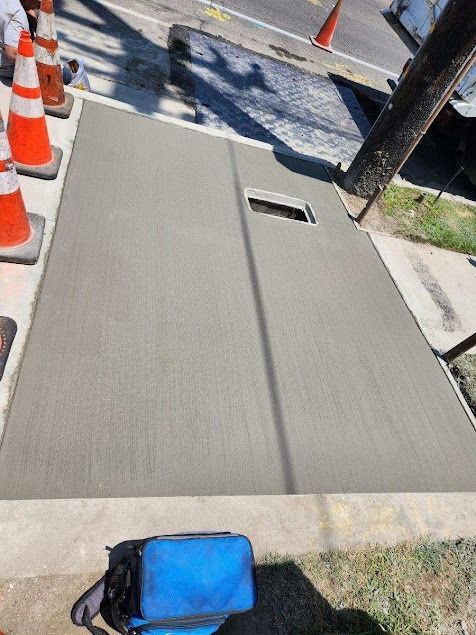 Freshly poured sidewalk concrete beside orange cones, with a blue bag in the foreground.