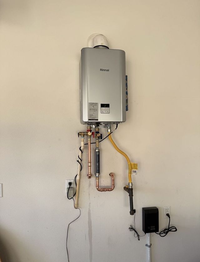 Wall-mounted gas water heater with exposed pipes, hoses, and a small control box on a plain wall