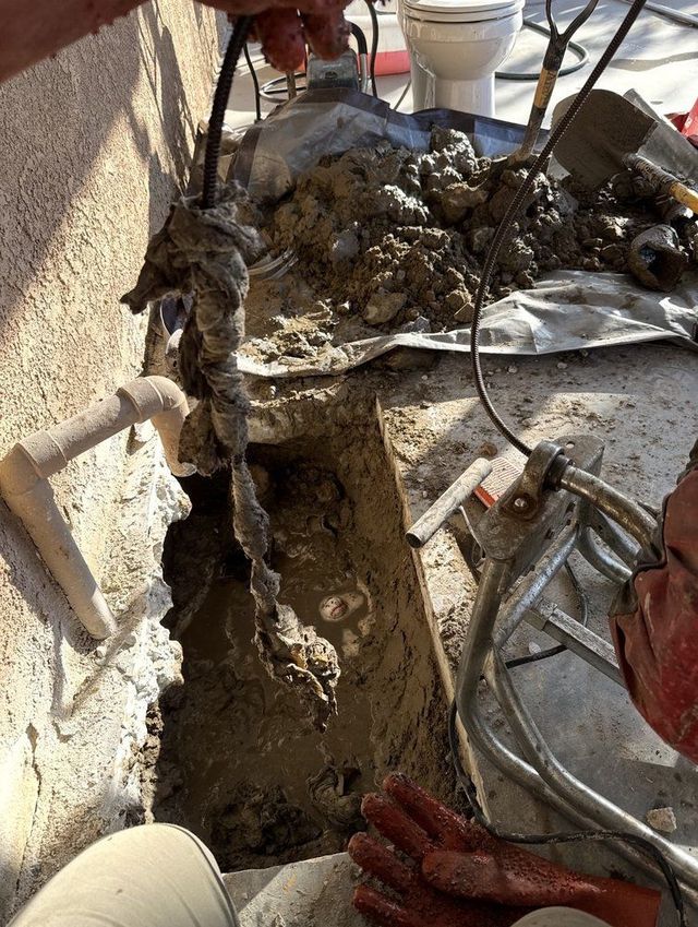 Worker digging a trench beside a wall, with muddy soil, tools, and construction debris nearby