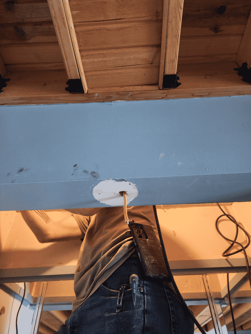 Person drilling into a painted gray beam, viewed from below. Wooden ceiling joists are above.