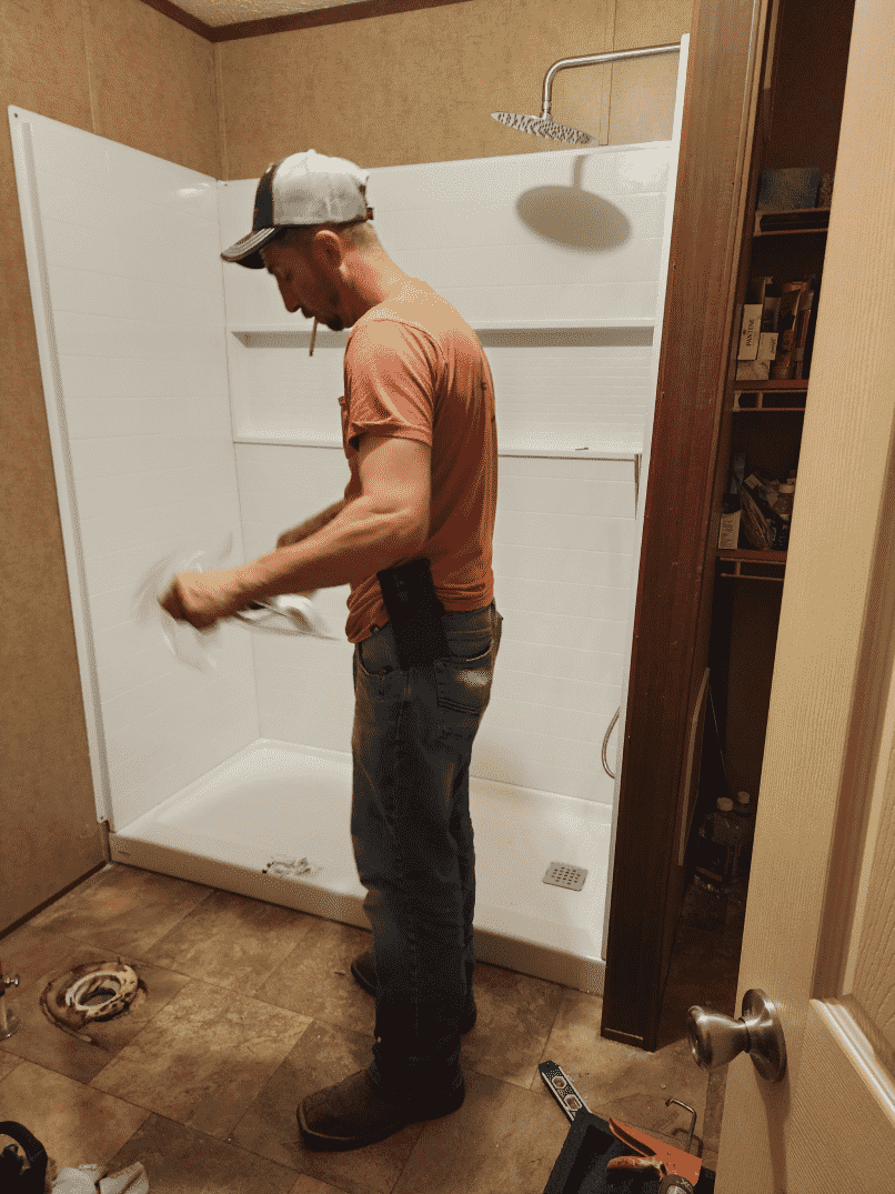 A person in a baseball cap works on installing a white shower stall in a bathroom.