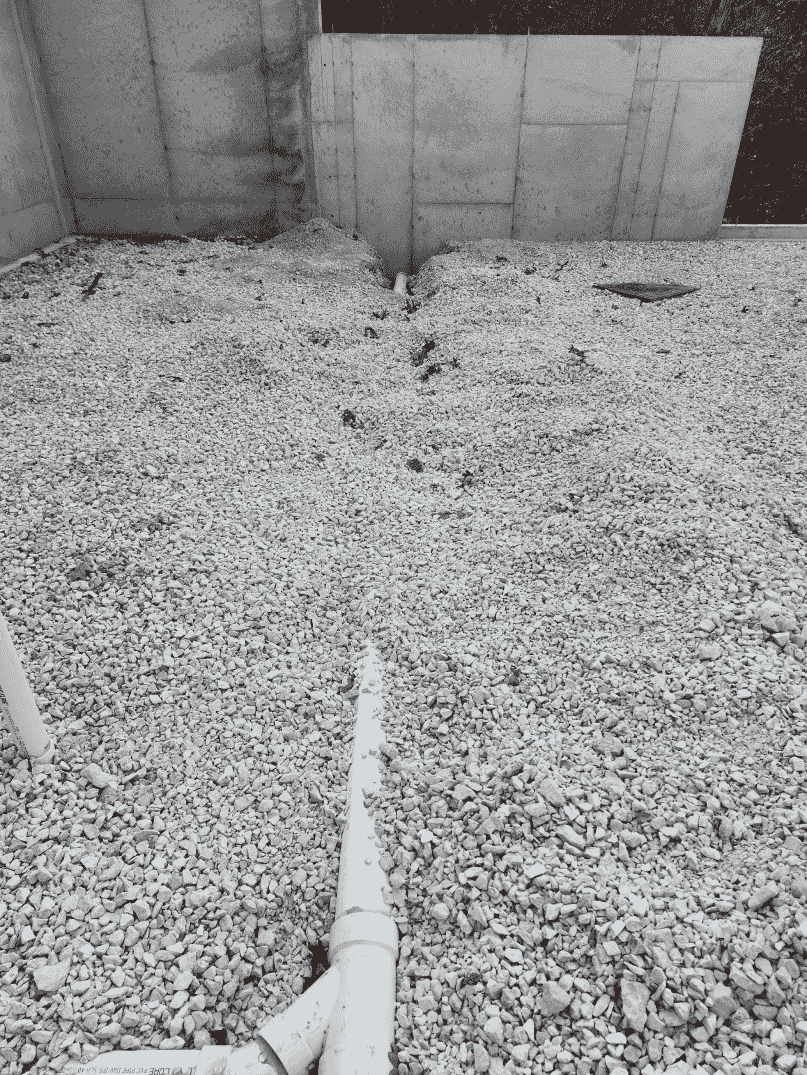 Gravel bed with exposed pipes, adjacent to a concrete foundation wall, viewed from a slightly elevated perspective.