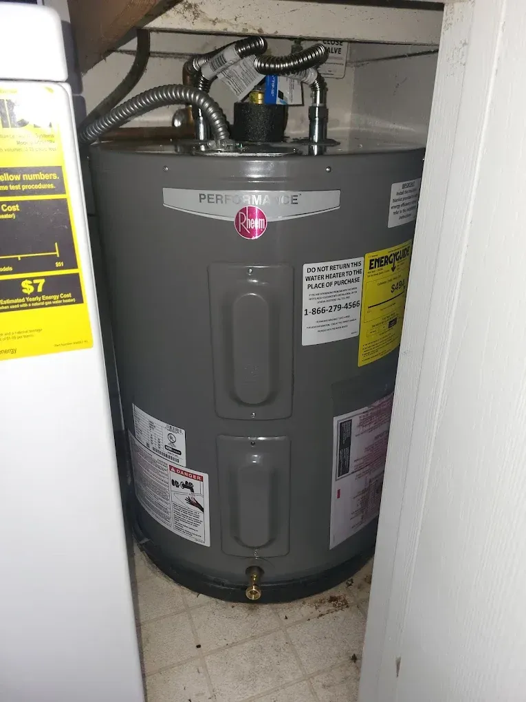 A gray Rheem electric water heater in a tight space, near a washing machine and a white wall.