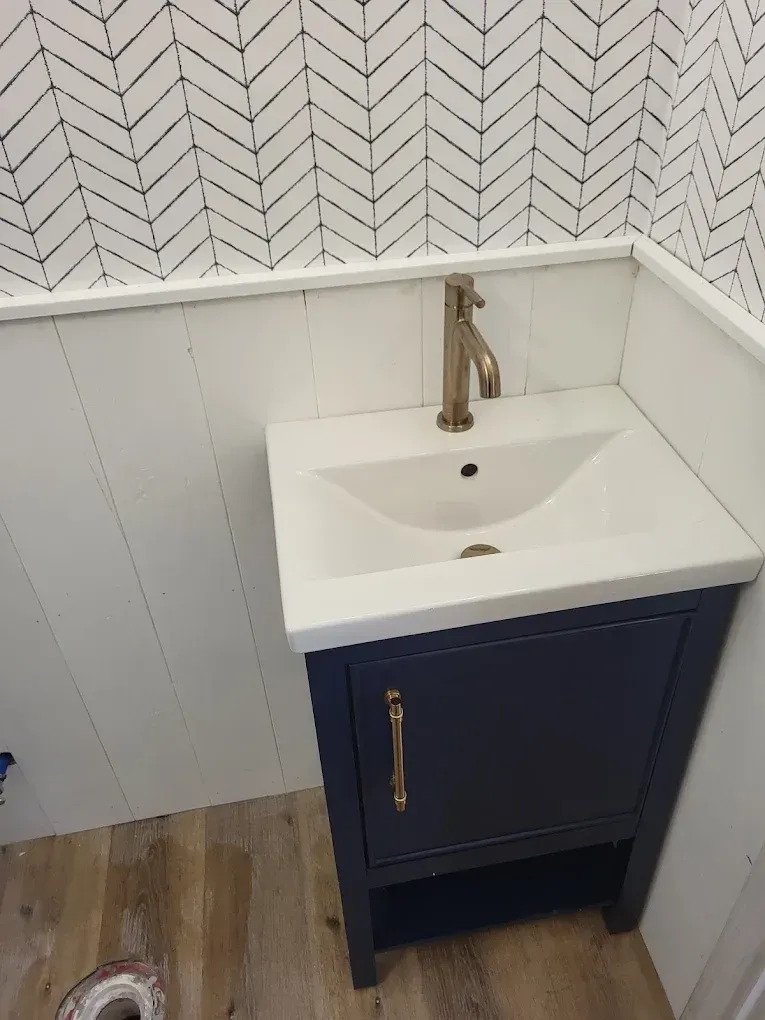 Small bathroom vanity with blue cabinet, white sink, and gold faucet, against chevron wallpaper.