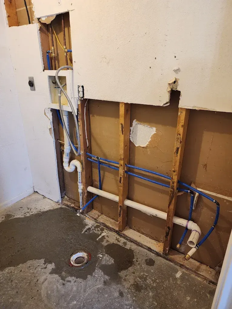 Bathroom wall under construction, with exposed framing, pipes, and concrete floor.