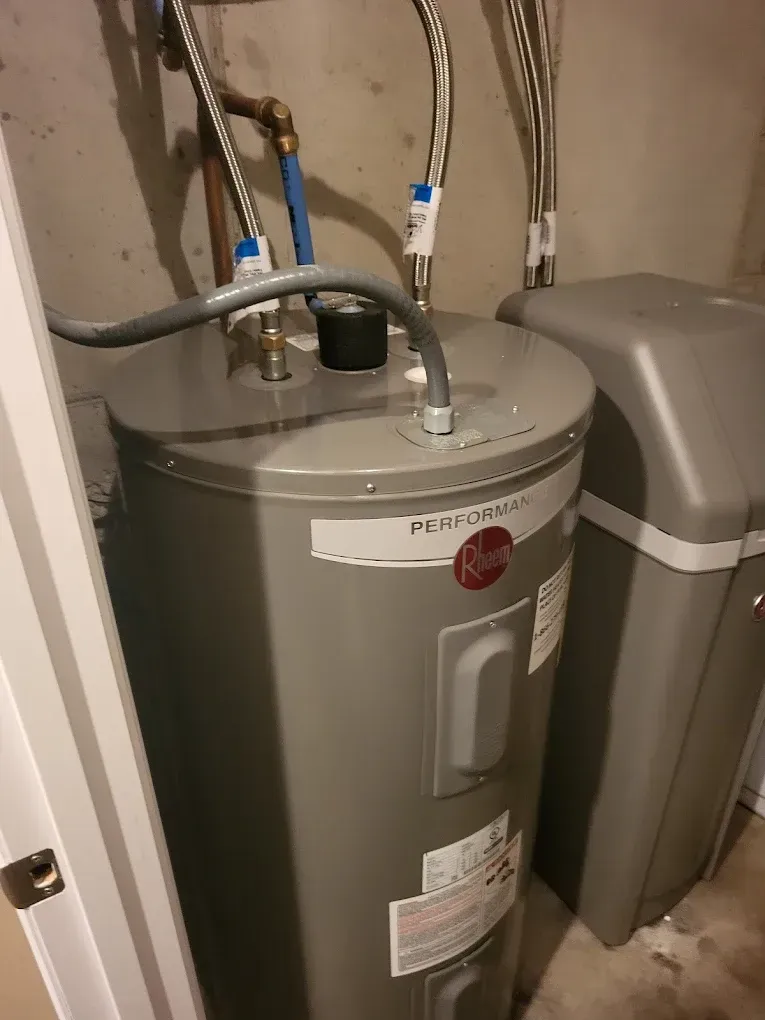 A gray Rheem water heater with pipes in a utility room next to a water softener.