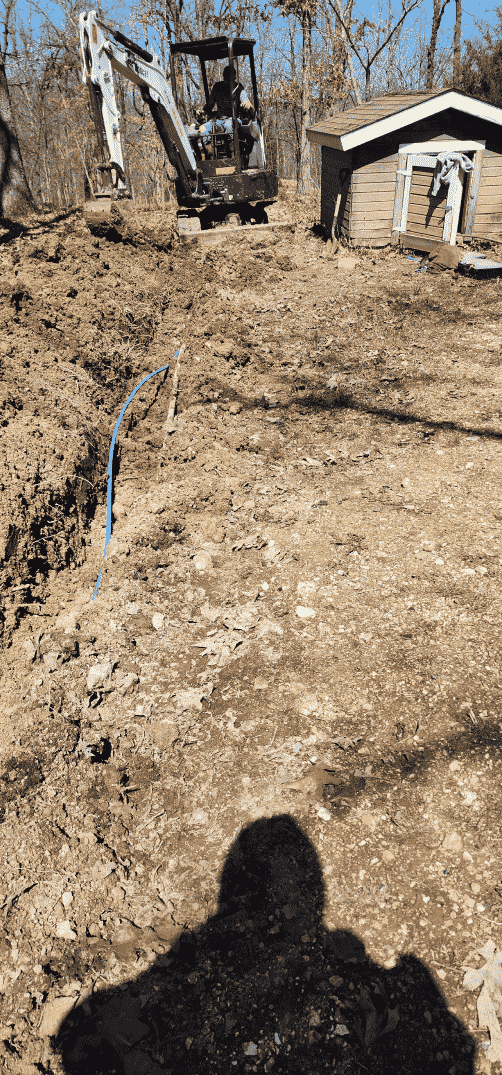 An excavator digging a trench near a small building in a dirt lot; shadow of a person in foreground.