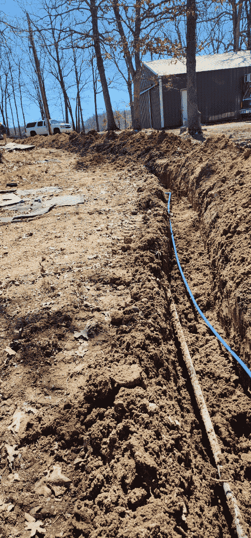 Trench dug in the dirt with a blue pipe and a brown pipe running through it, building in the background.