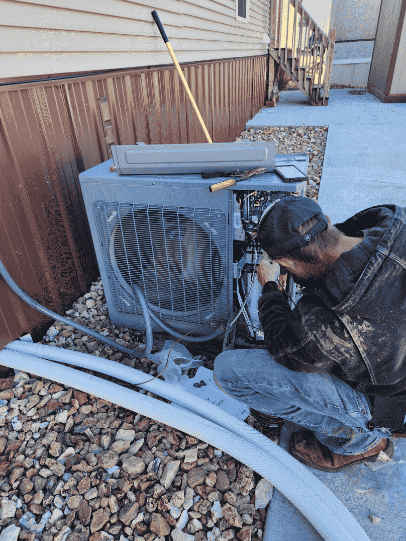A person repairs an outdoor air conditioning unit next to a building.